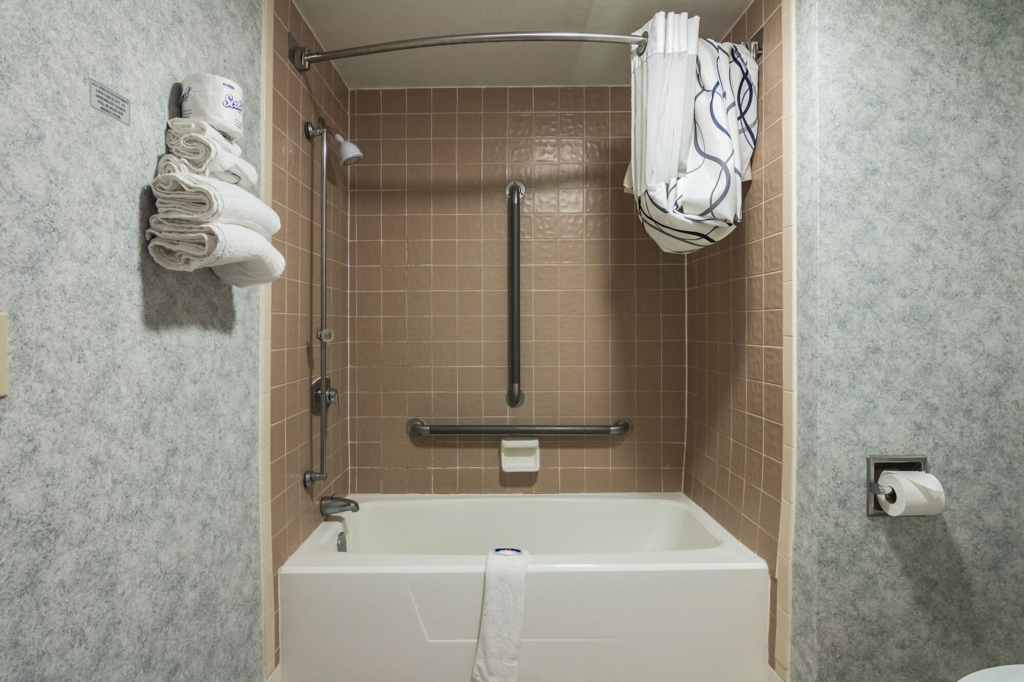 A bathroom with a bathtub, a shower, and a handrail. There are towels and a toilet paper holder visible.