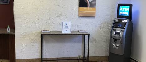 A clean, well-lit room with a tiled floor, a small table with a sign, an ATM machine, and a poster on the wall.