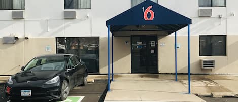 Un edificio con toldo azul marcado con el número 6, que indica un hotel o motel. Hay un estacionamiento con estación de carga para Tesla.