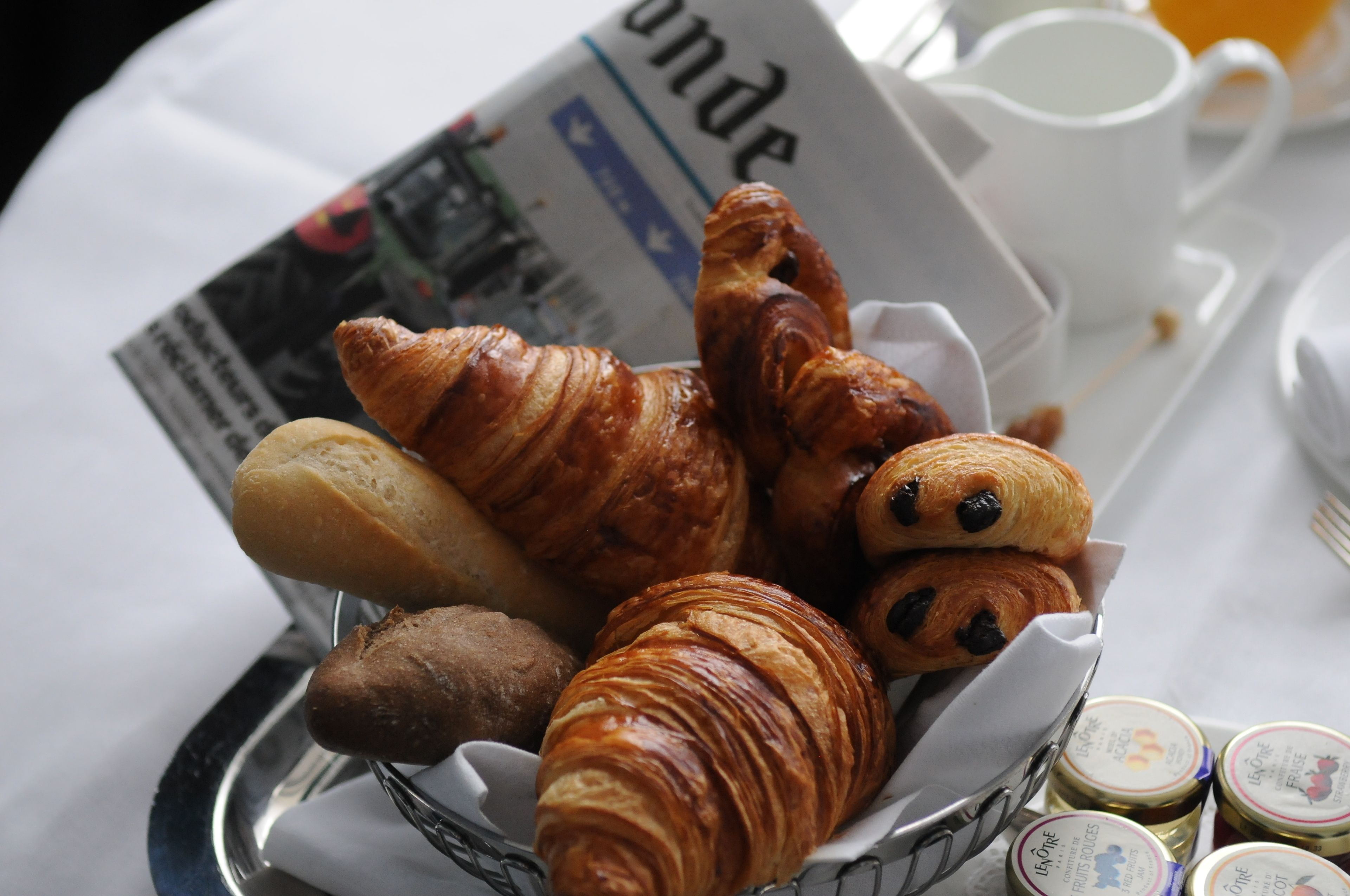 A tray of assorted pastries including croissants and a baguette, accompanied by a newspaper and a white cup.