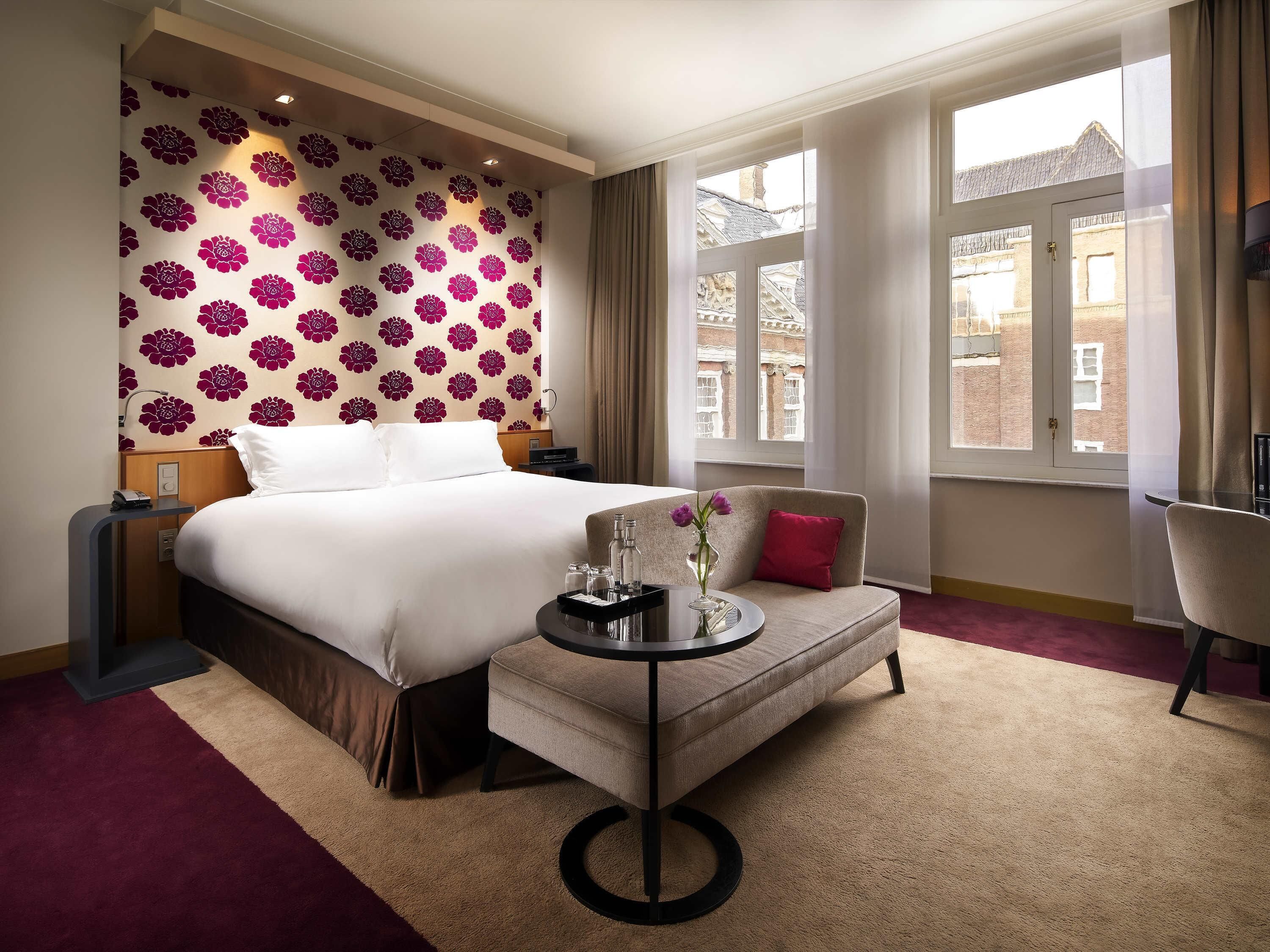 Ett hotellrum med en stor säng, en liten soffa, ett runt bord med en vas och utsikt över byggnader genom fönstret.