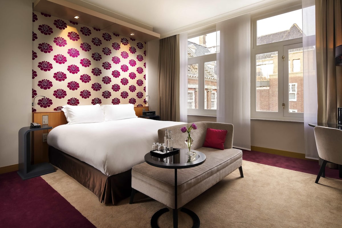 A hotel room with a large bed, a small sofa, a round table with a vase, and a view of buildings through the window.