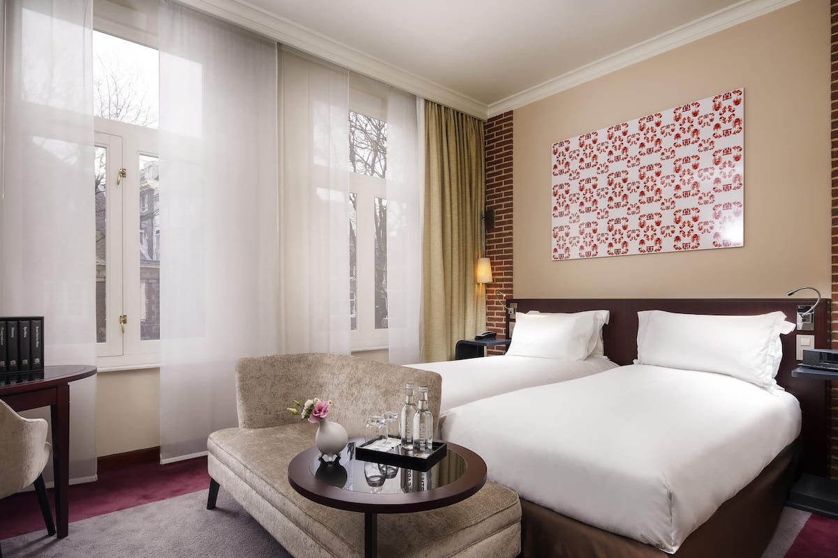 A hotel room with a bed, a chair, a small table with a vase, and a wall with a floral pattern.