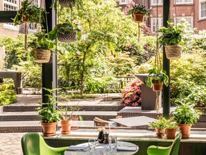 Uma área externa com assentos, vasos de terracota e cestos suspensos com vegetação, uma mesa com cadeiras e vista para um edifício de tijolos e um jardim exuberante.