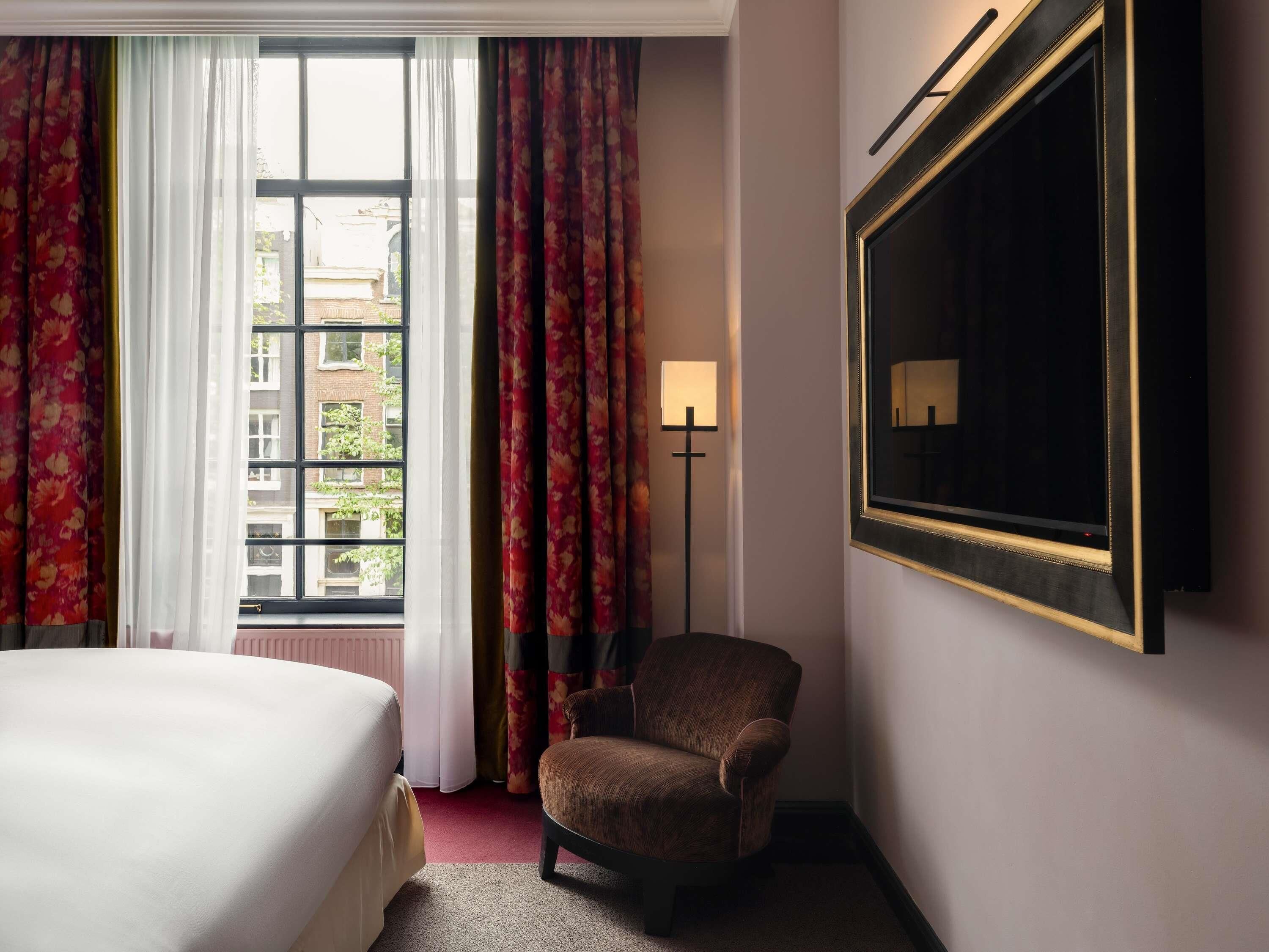 Una camera d'albergo con un letto, una sedia, un'ampia finestra con tende, una lampada da terra e uno specchio a parete.