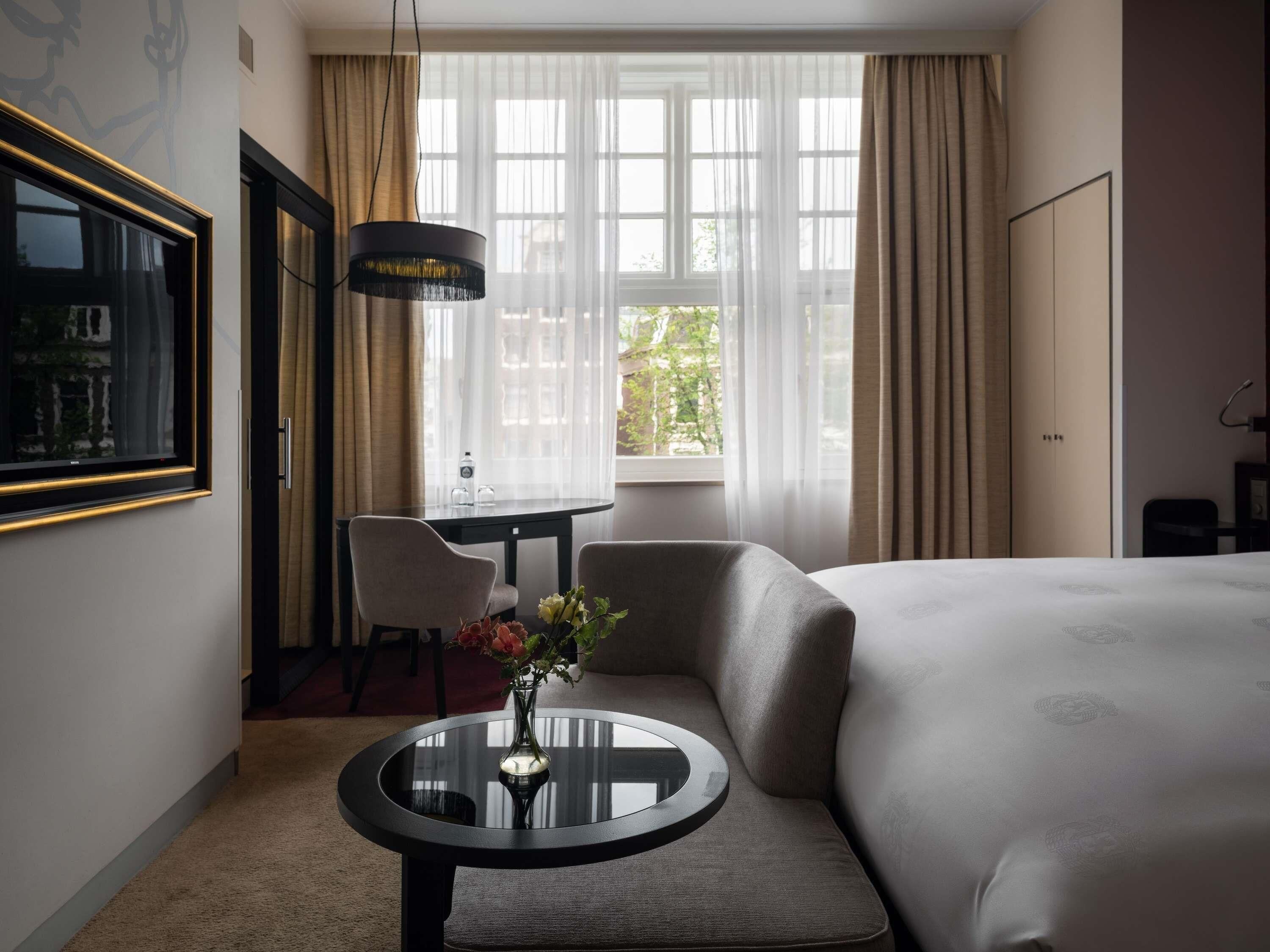 A hotel room with a bed, a round table with a vase, a chair, a desk, and a window with curtains.