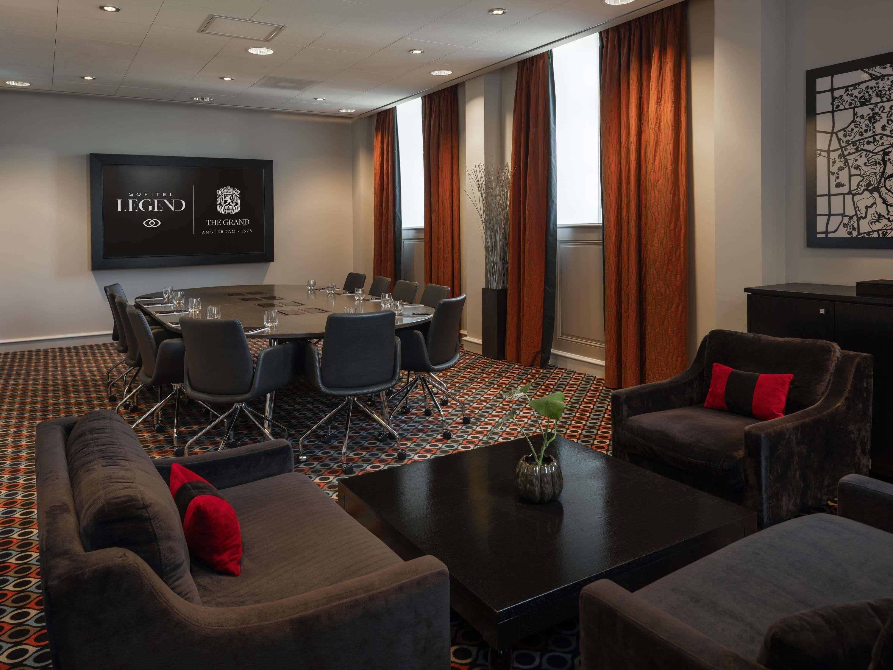 A conference room with a large table, chairs, a sofa, and a TV screen displaying 'SOFITEL LEGEND THE GRAND AMSTERDAM 1578'.