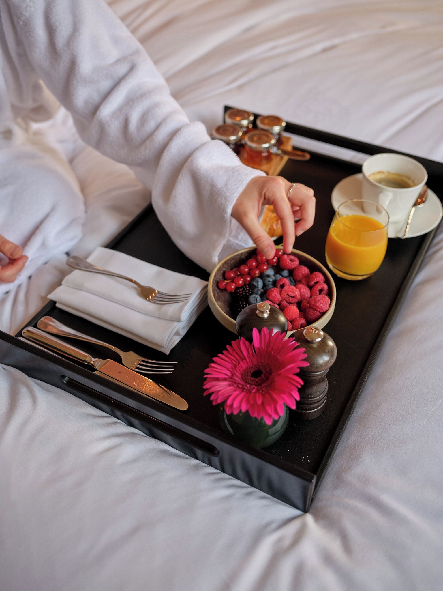 Un plateau contenant des aliments pour le petit-déjeuner, notamment des fruits rouges, un verre de jus d’orange et une tasse de café, posé sur un lit.