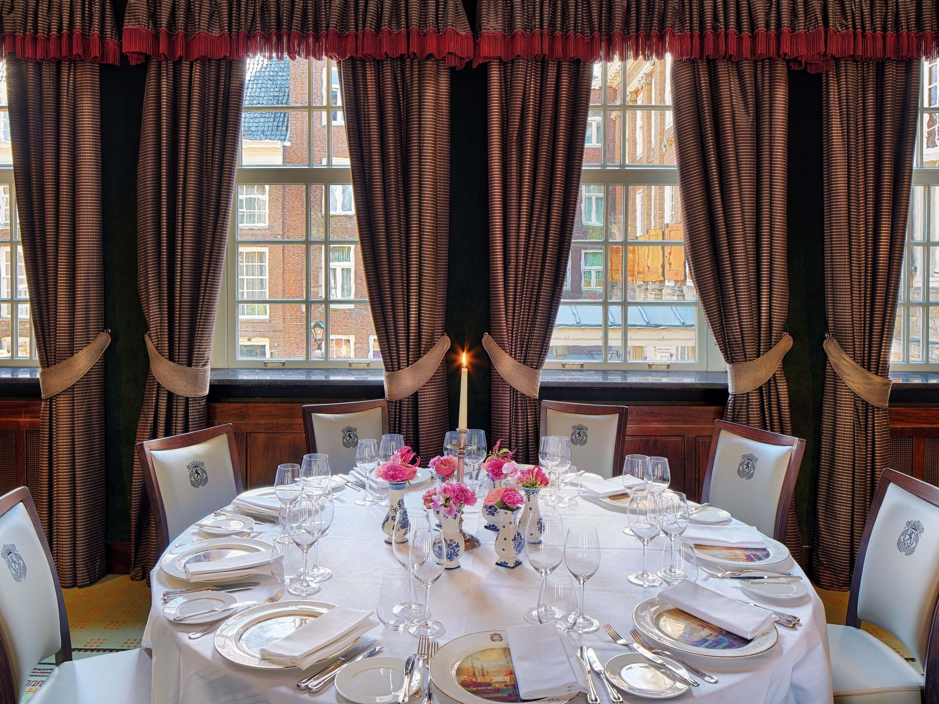 A dining room with a round table set for six, flanked by curtains and a window with a view of buildings.