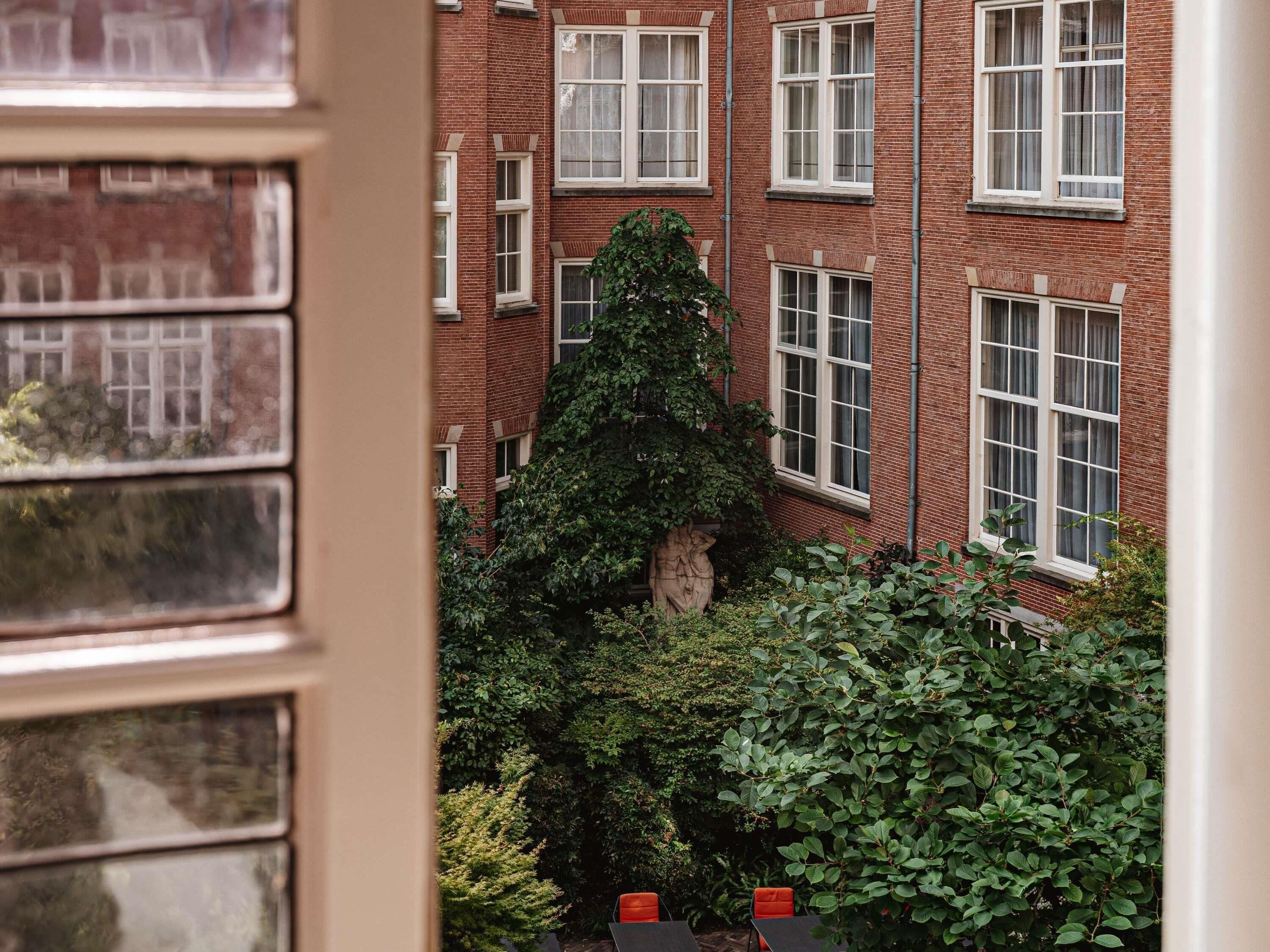 A red brick building with multiple windows and a garden area with greenery and a stone sculpture.