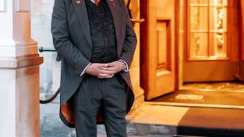 A man in a dark suit standing in front of a wooden door with a glass panel.