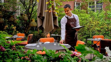 Un camarero con delantal, de pie junto a una mesa con un menú, rodeado de mesas al aire libre y vegetación.