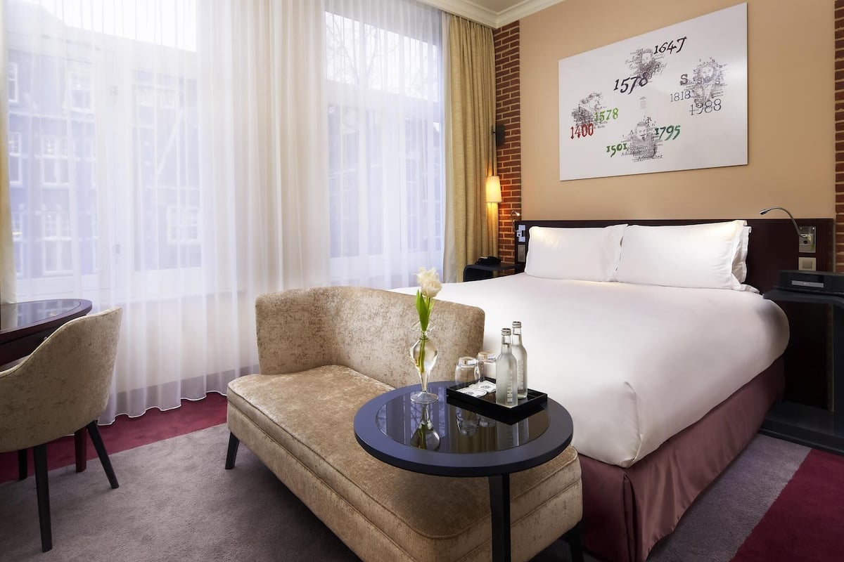 A hotel room with a bed, a chair, a small table with a vase and bottles, and a wall with a historical timeline.
