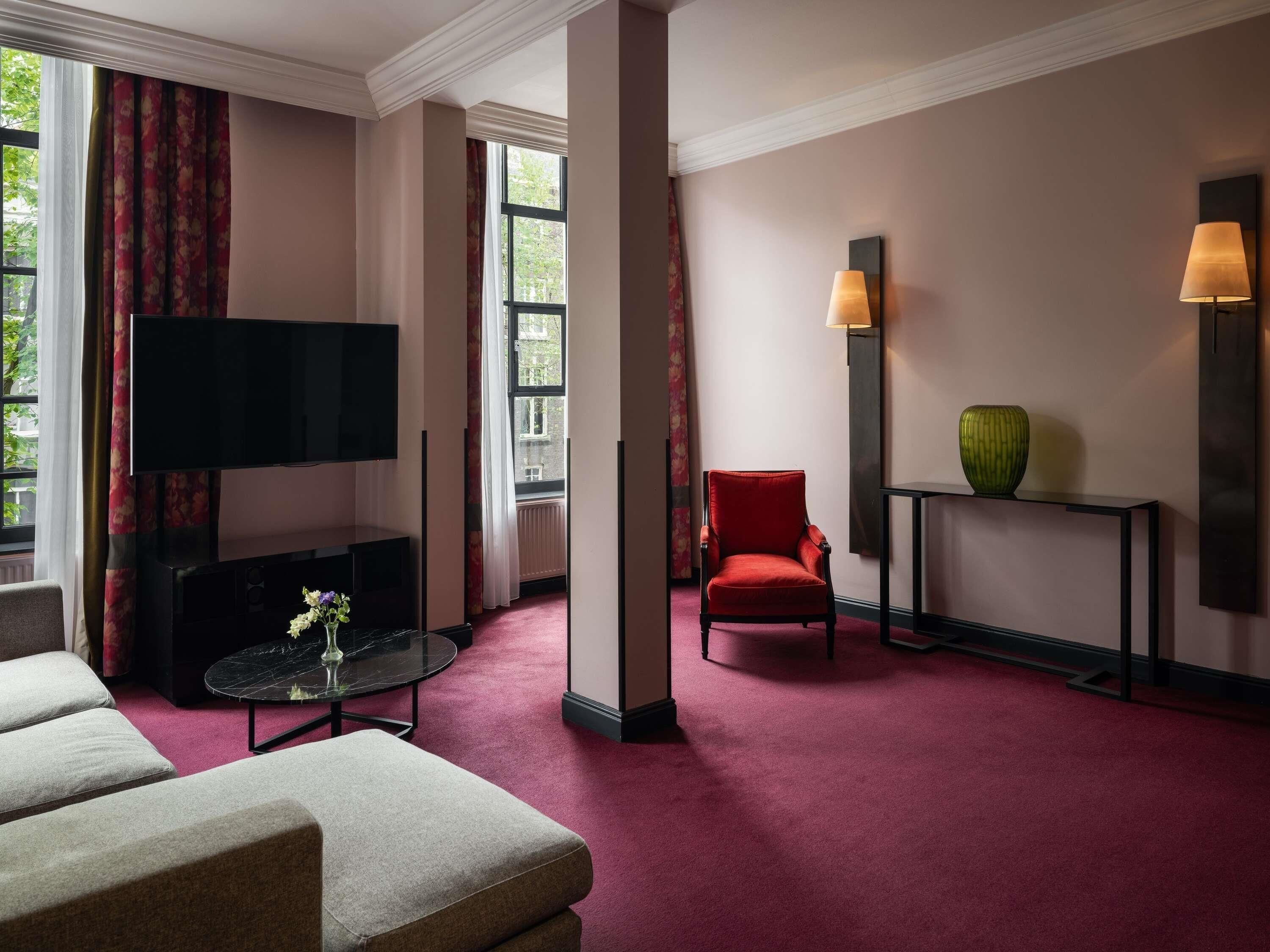 Une chambre d’hôtel équipée d’un téléviseur à écran plat, d’un fauteuil rouge, d’une petite table avec un vase et d’une console sur laquelle est posé un vase.