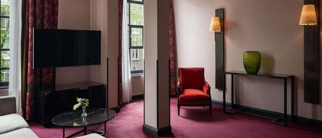 Habitación de hotel con televisor de pantalla plana, un sillón rojo, una mesita con un florero y una consola con otro florero.