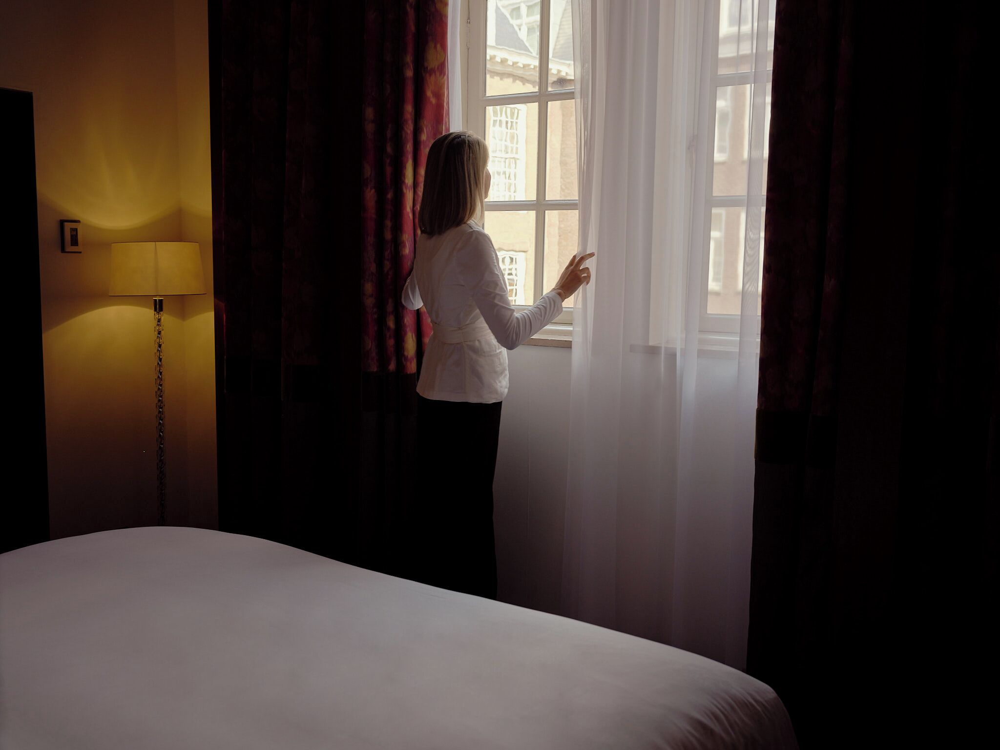 Quarto de hotel com cama, abajur e janela com cortinas.