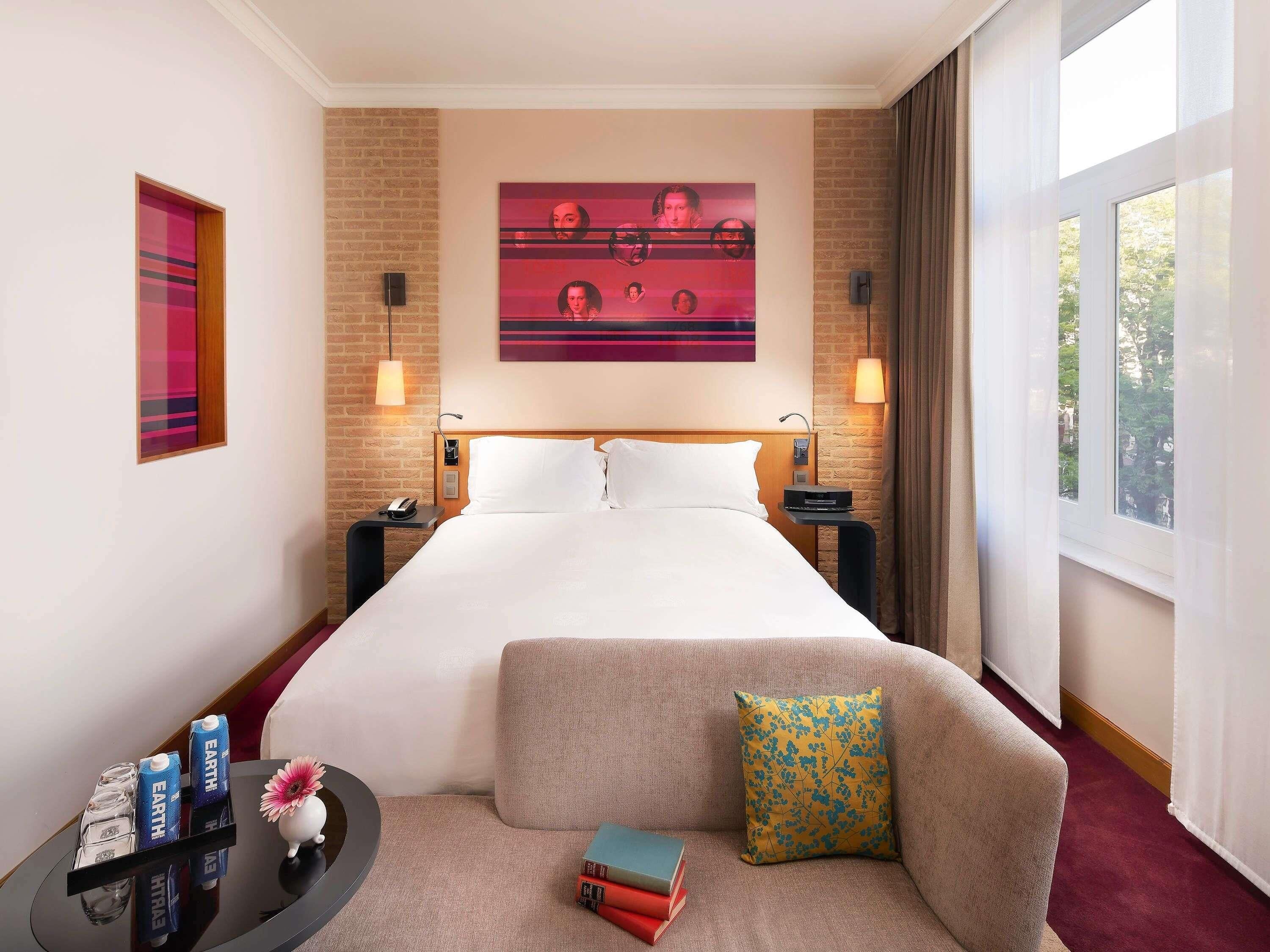Habitación de hotel con cama, mesitas de noche, un pequeño sofá, una mesa de centro y un ventanal grande.