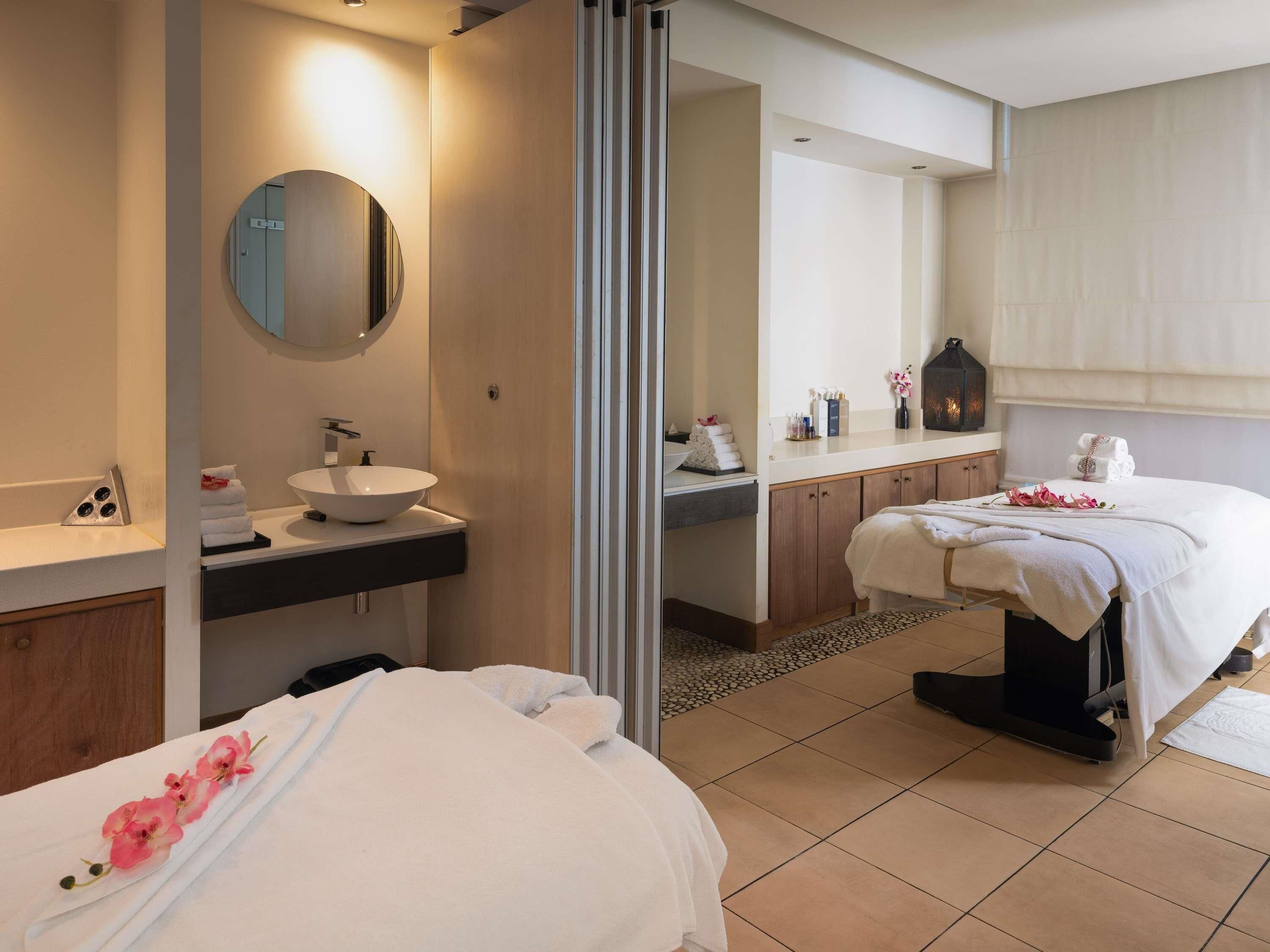 A modern hotel room with a large bed, a separate massage table, a round mirror, a sink, and a vanity with toiletries.