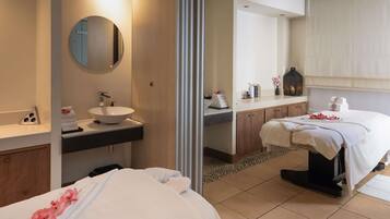 A modern hotel room with a large bed, a separate massage table, a round mirror, a sink, and a vanity with toiletries.