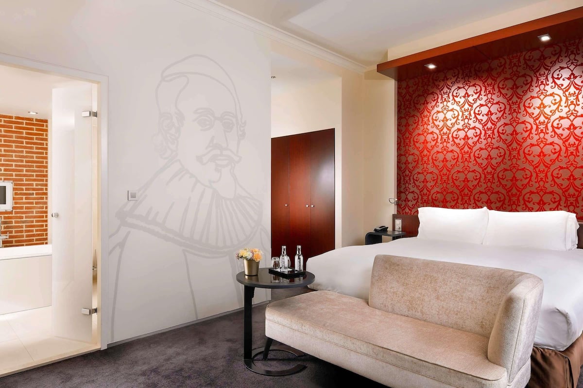 A hotel room with a bed, a small sofa, a nightstand, a wall with a red patterned design, a door, and a wall with a white design.
