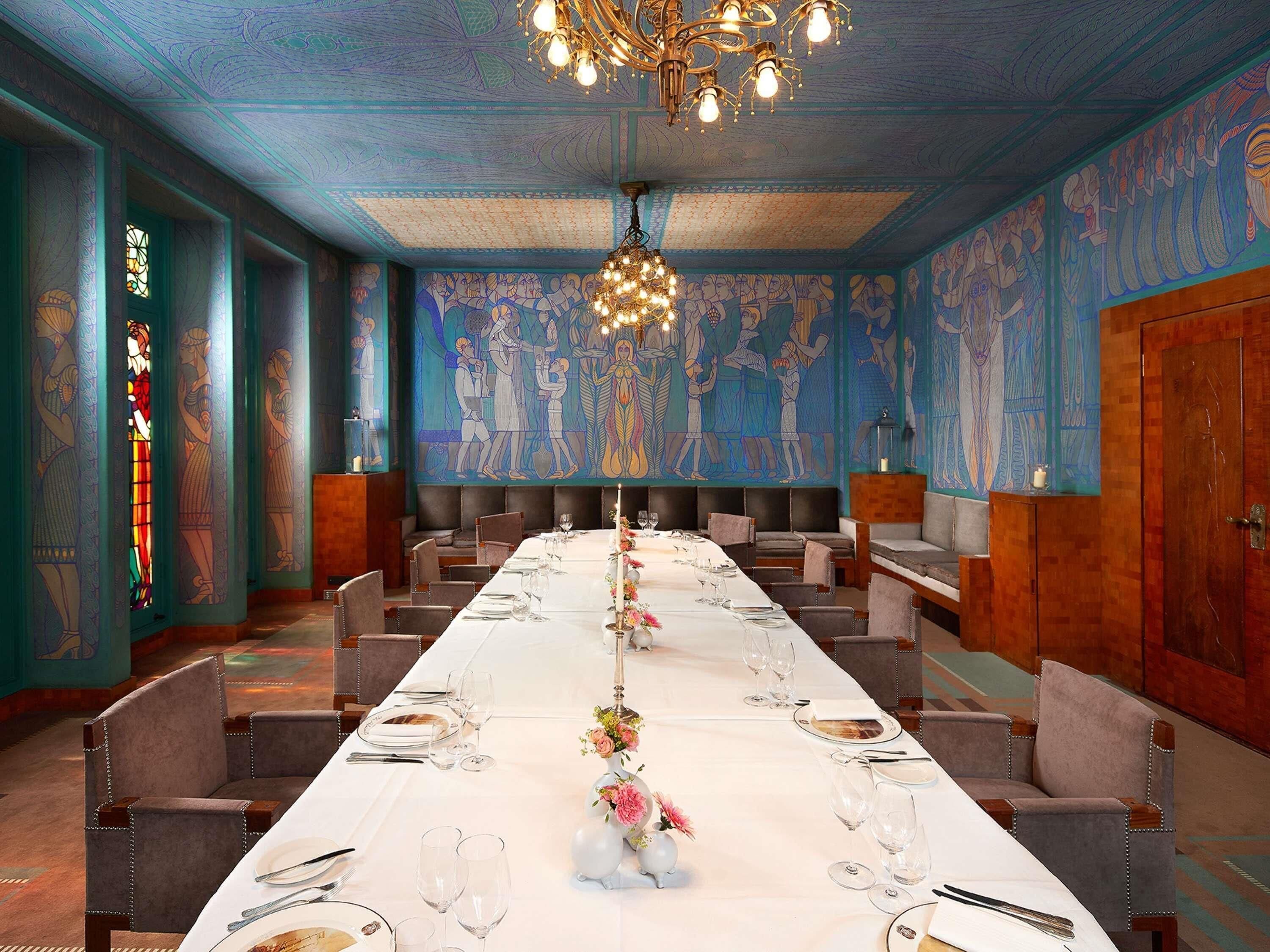 Una larga mesa de comedor con sillas, una araña y un mural en las paredes.