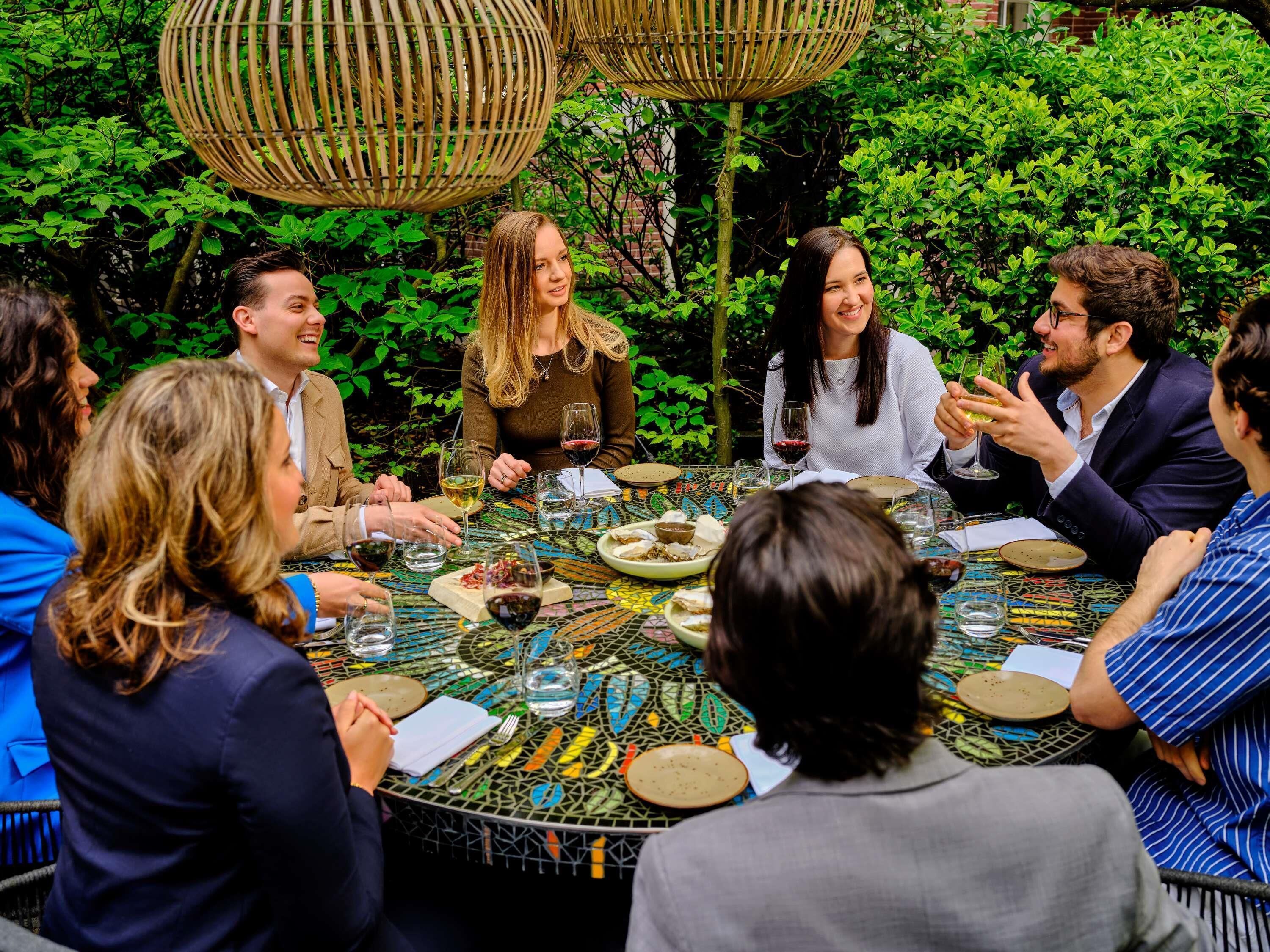 A group of people seated around a round table with a mosaic tablecloth, engaged in conversation.