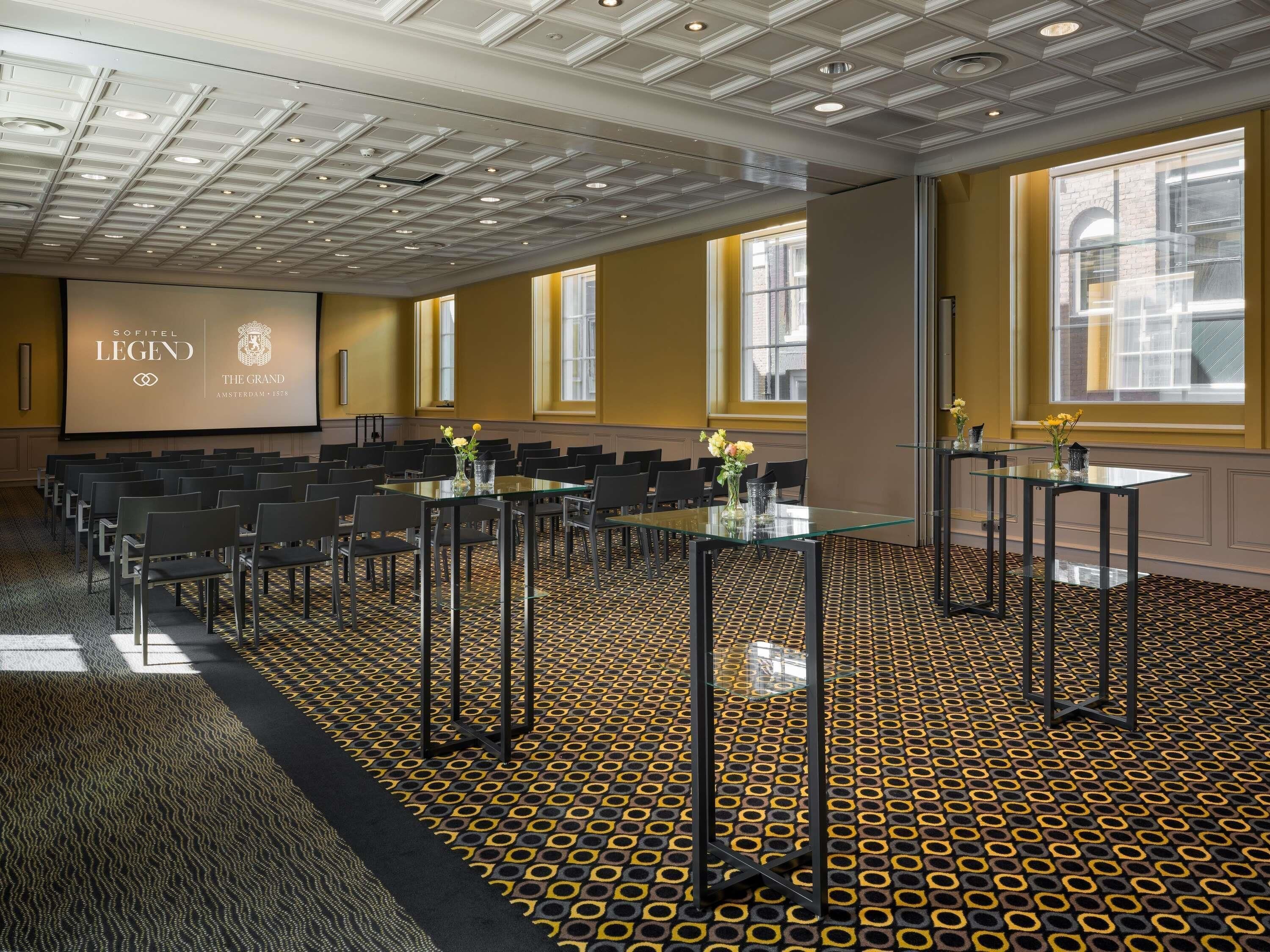 Une salle de conférence dotée d’un tapis à motifs, d’un grand écran affichant le logo de Sofitel Legend, et de plusieurs tables entourées de chaises.