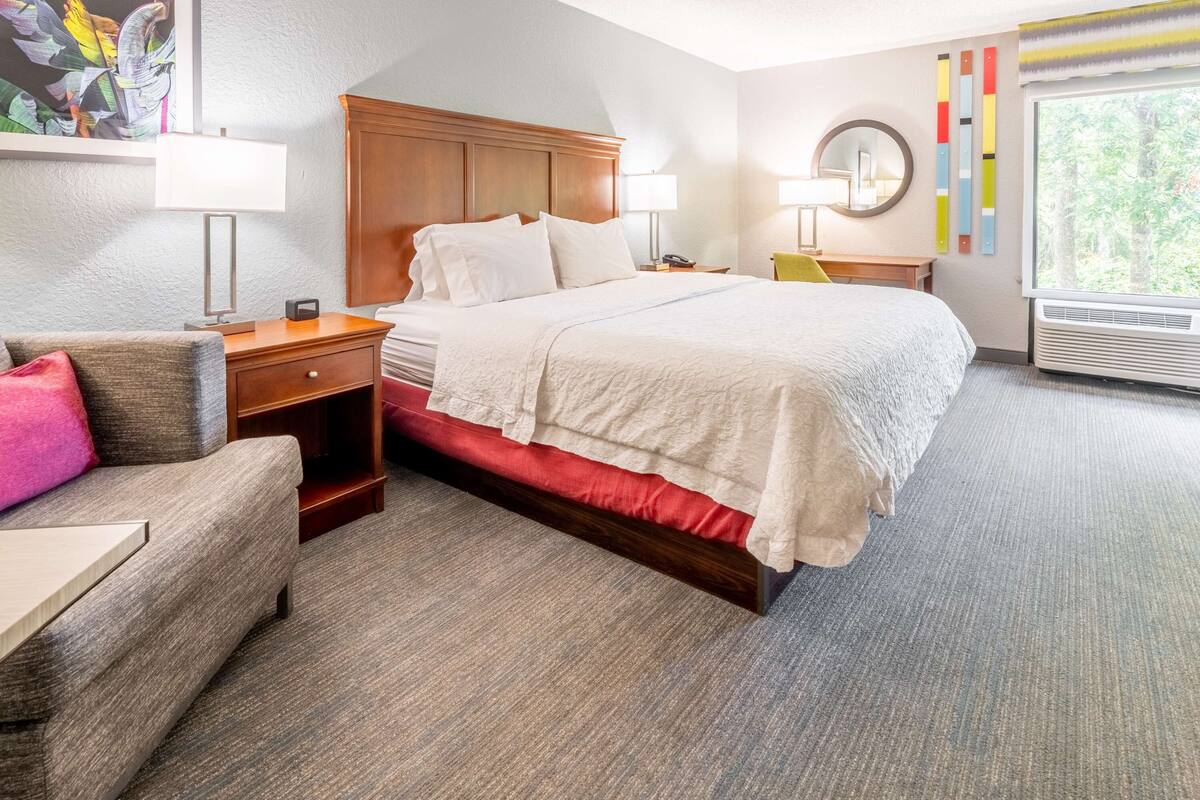 A hotel room with a bed, bedside tables, a chair, a painting, a mirror, and a window with a view of trees.