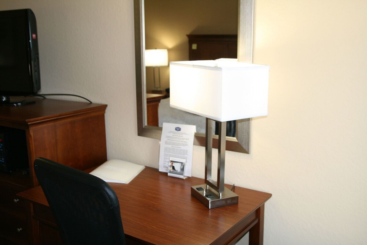 Habitación de hotel con un escritorio de madera, una lámpara, un espejo, un televisor y un documento sobre el escritorio.