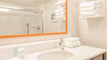 Un baño de hotel con un espejo grande, un lavamanos y una ducha. Hay toallas en un estante y una pequeña foto en un marco en la pared.