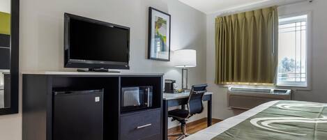 A hotel room with a bed, a desk with a computer, a television, a refrigerator, a lamp, and a window with curtains.