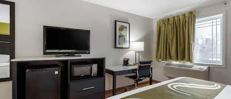 Habitación de hotel con cama, escritorio con silla, televisión, microondas, refrigerador pequeño, ventana con cortinas y aire acondicionado de pared.