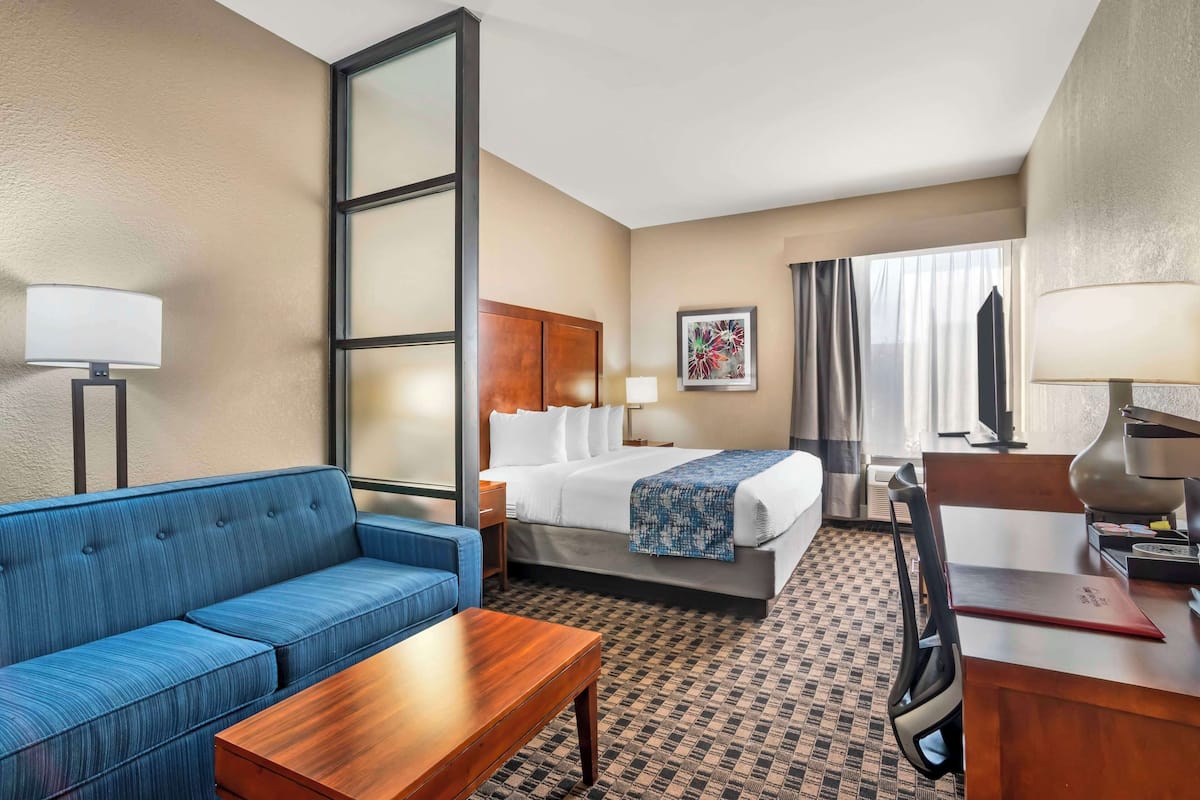 Une chambre d’hôtel avec un lit, un canapé bleu, une table basse en bois, un bureau avec une lampe et une grande fenêtre avec des rideaux.