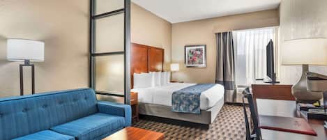 A hotel room with a bed, a blue sofa, a wooden coffee table, a desk with a lamp, and a large window with curtains.