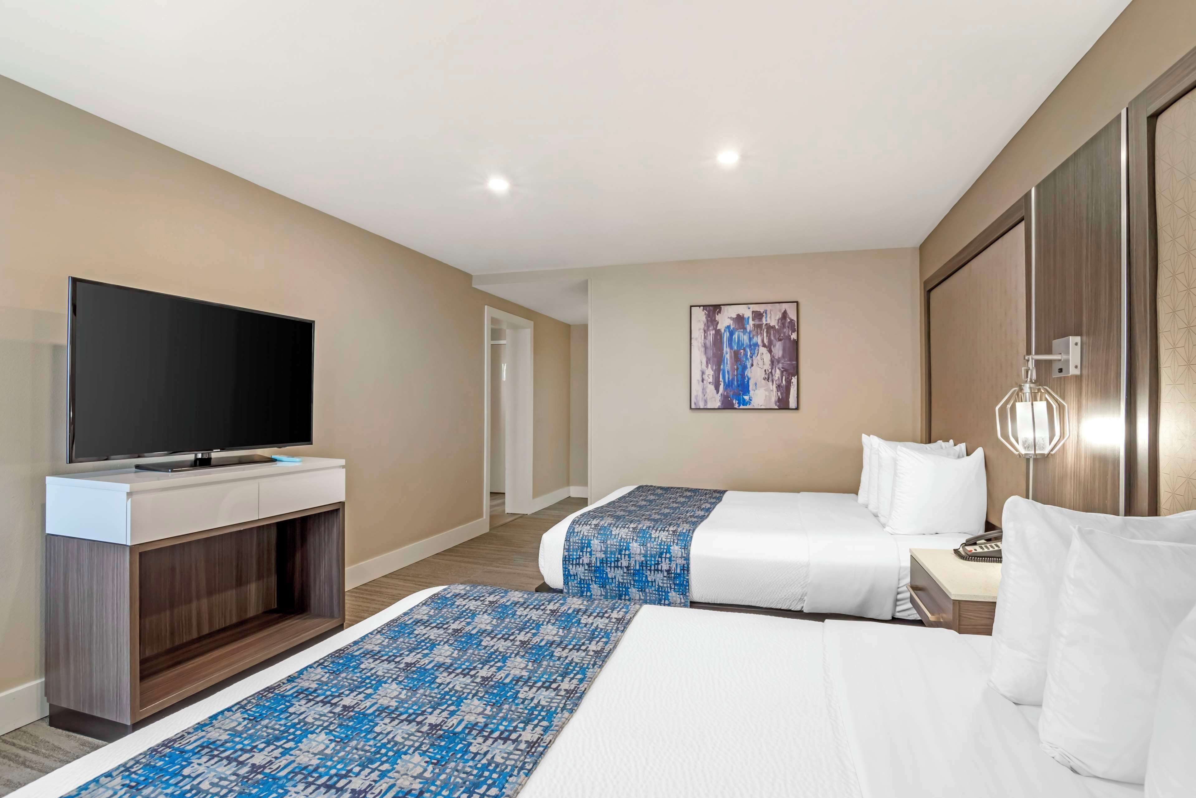 Une chambre d’hôtel équipée d’un téléviseur à écran plat, de deux lits avec du linge de lit blanc, d’un tapis à motifs et d’un tableau accroché au mur.