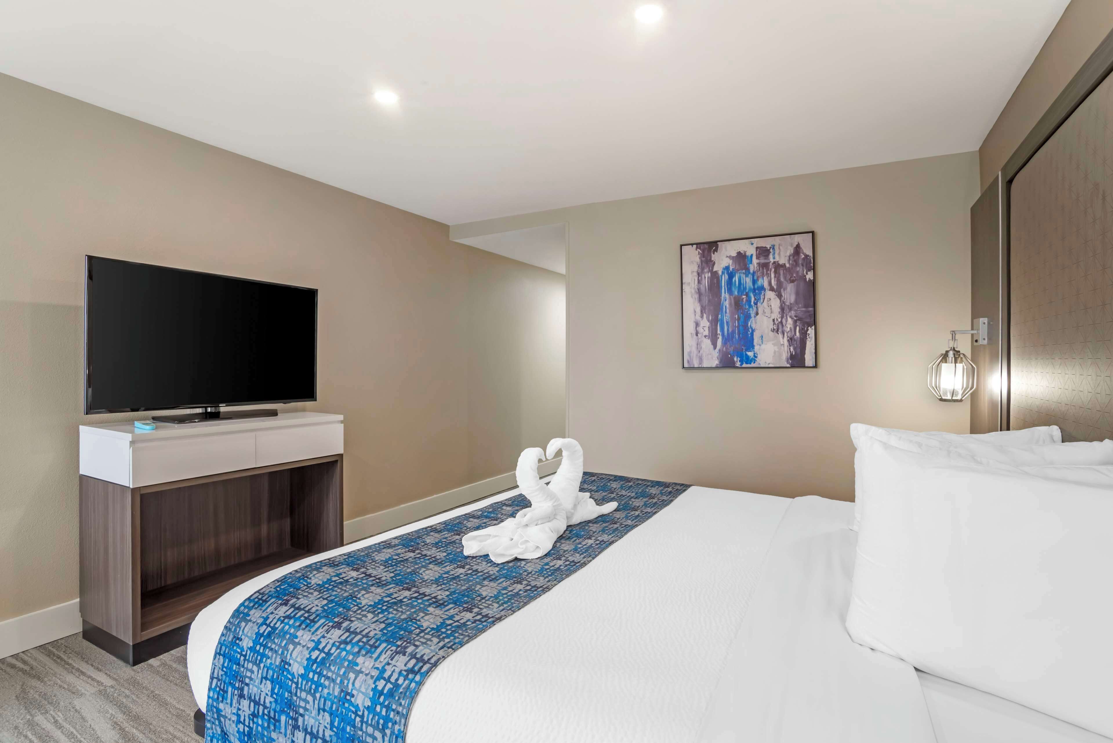 A hotel room with a bed, a flat-screen TV, a painting on the wall, and a wall-mounted lamp.