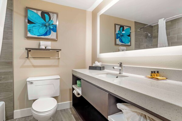 A modern bathroom with a toilet, sink, and shower. There are two framed floral artworks on the wall, a towel rack, and a shelf with toiletries.
