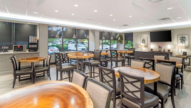 Interior moderno de restaurante con mesas y sillas redondas, una zona de cocina y grandes ventanales con vistas a coches aparcados afuera.
