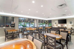 Interior moderno de restaurante con mesas y sillas redondas, una zona de cocina y grandes ventanales con vistas a coches aparcados afuera.