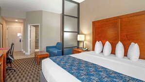 Una habitación de hotel con una cama grande, un escritorio con una silla, un sillón azul, una mesita de noche de madera con una lámpara, y un armario con espejo.