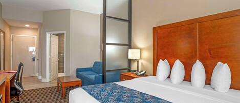 Ein Hotelzimmer mit einem großen Bett, einem Schreibtisch mit Stuhl, einem blauen Sessel, einem Nachttisch aus Holz mit Lampe und einem Kleiderschrank mit Spiegel.