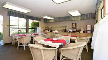 A dining area with wicker chairs and tables, a kitchen area with a coffee machine, and a window with a view of greenery.