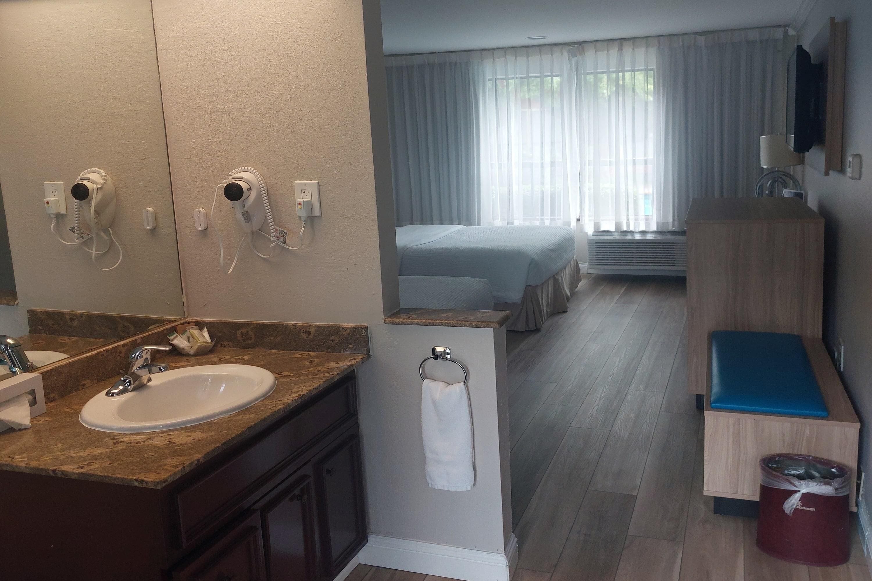 Une salle de bain d’hôtel avec un lavabo, un miroir, un porte-serviettes, et un lit visible à travers la fenêtre.