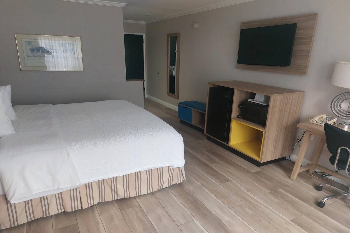 A hotel room with a large bed, a wardrobe, a desk with a chair, a television, and a framed picture on the wall.