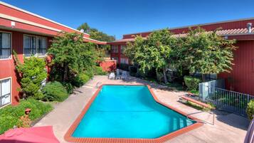A swimming pool surrounded by red buildings with windows, trees, and a clear blue sky.