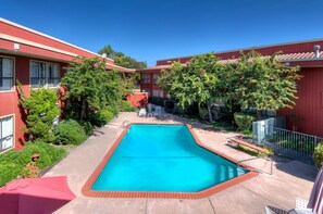 A swimming pool surrounded by red buildings with windows, trees, and a clear blue sky.