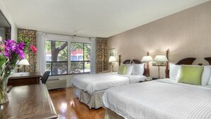 A hotel room with two beds, a desk, a chair, and a window with a view of trees and buildings.