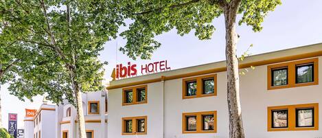 Ein Hotel namens „ibis HOTEL“ mit heller Fassade, zahlreichen Fenstern und einem umlaufenden Garten mit Blumen.