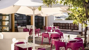 An outdoor seating area with white and purple chairs and tables, covered by large white umbrellas, and surrounded by greenery and a cobblestone path. - ibis Evora Hotel (Évora)