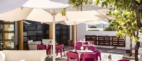 An outdoor seating area with white and purple chairs and tables, covered by large white umbrellas, and surrounded by greenery and a cobblestone path.